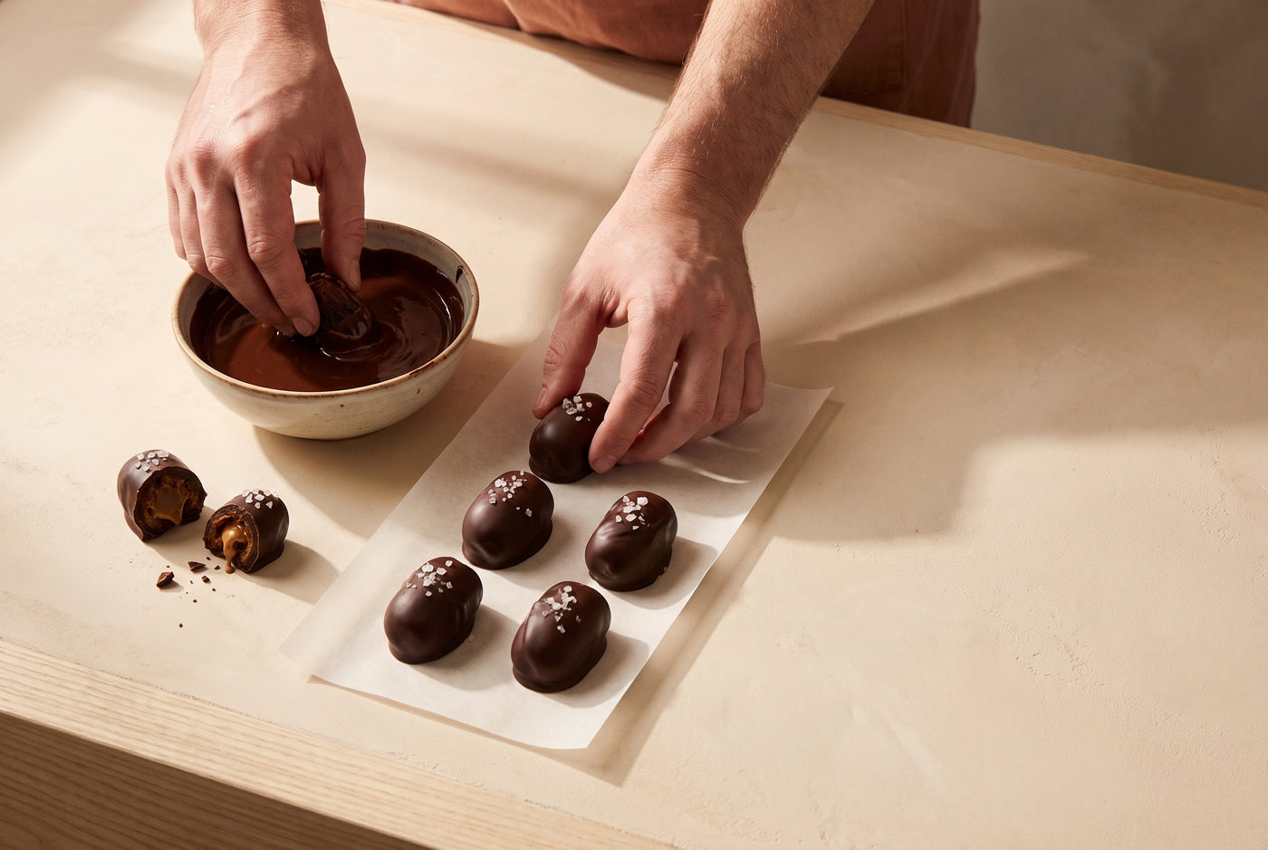 Hands dipping a Medjool date into melted dark chocolate and setting finished dates on parchment